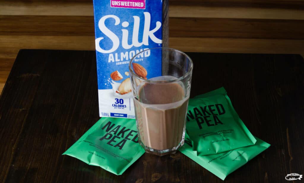 A class of naked pea protein powder, surrounded by packages of the powder and a carton of almond milk