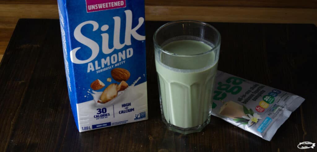 A glass of Vega protein shake with the vega package to one side and a carton of almond milk to the other side.