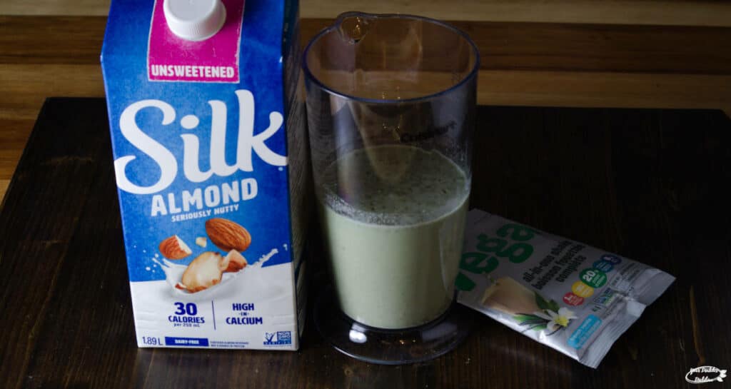 A shake glass full of a Vega protein shake, with a carton of almond milk beside it
